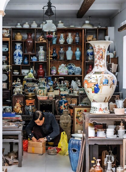 A cluttered room filled with various pottery and ceramics, a large ornate vase, and a person sorting through items. 4