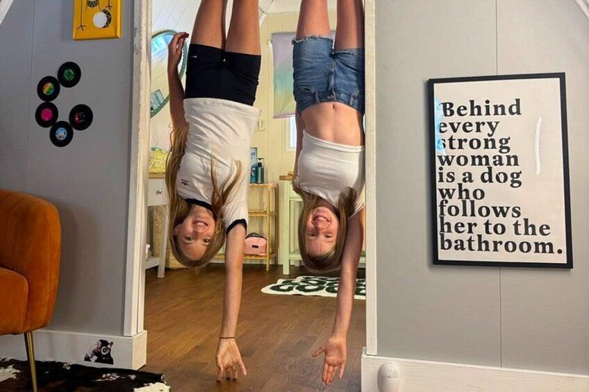 Dos personas haciendo paradas de manos en una habitación con un cartel en la pared que dice 'Behind every strong woman is a dog who follows her to the bathroom'. 3
