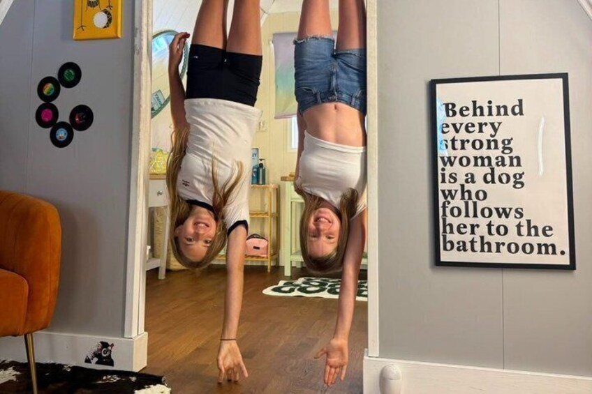 Two people doing handstands in a room with a wall sign that reads 'Behind every strong woman is a dog who follows her to the bathroom.' 2