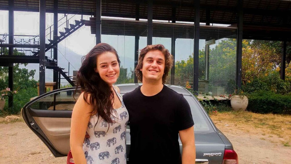 Two people standing in front of a car with a covered structure and greenery in the background. 2