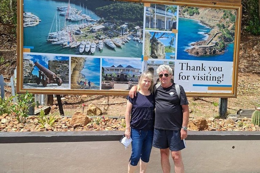 A couple posing in front of a large informational sign with multiple photographs of coastal scenes and a message thanking visitors. 2