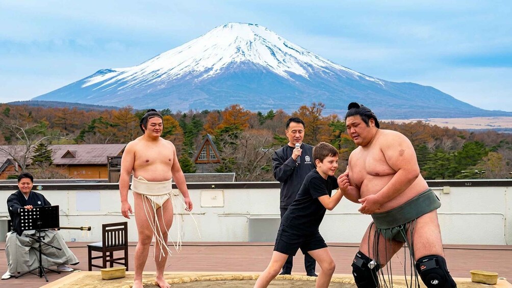 Four sumo wrestlers, a man with a microphone, and a child on a rooftop with a snow-capped mountain in the background. 5
