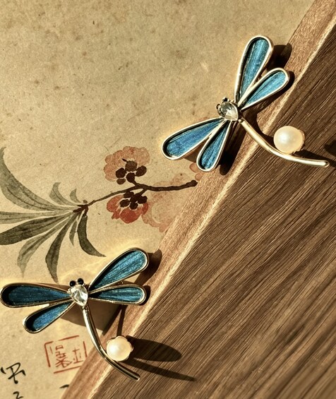Two dragonfly earrings with blue wings and pearl accents, resting on a wooden surface with floral patterns. 2
