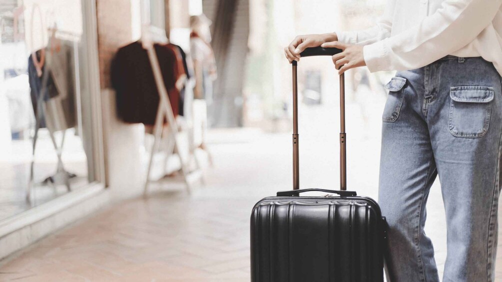 A person with a black suitcase, wearing a white shirt and blue jeans, standing in a corridor with a blurred background. 2