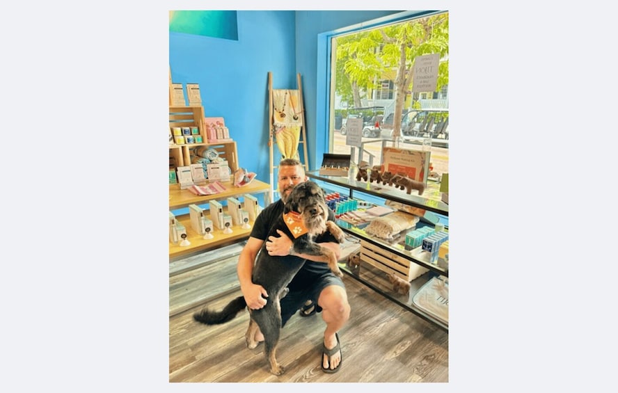 A person sitting on a chair in a room with a wooden floor, holding a dog. There are shelves with various items and a window with a view of the outside. 2