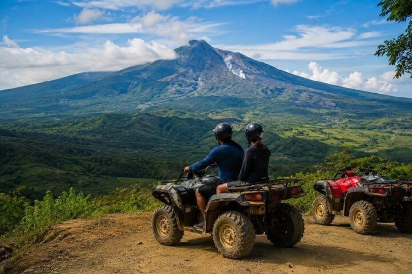 Two people on an ATV, a volcano, and a forested landscape. 1