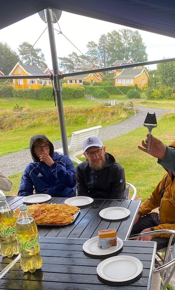 Three people sitting at a table with food and drinks, outdoors with houses and greenery in the background. 2