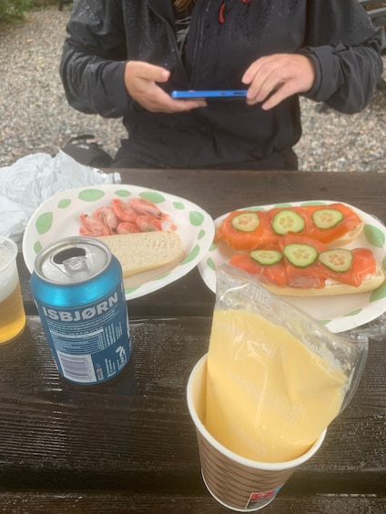 A person holding a blue smartphone, a can of ISBJÖRN beer, a sandwich with shrimp and cucumber slices, and a cup of cheese. 5