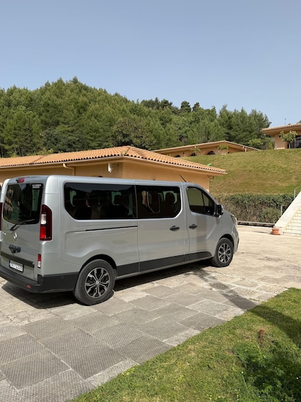 A silver van parked on a paved area with a hillside and buildings in the background. 3
