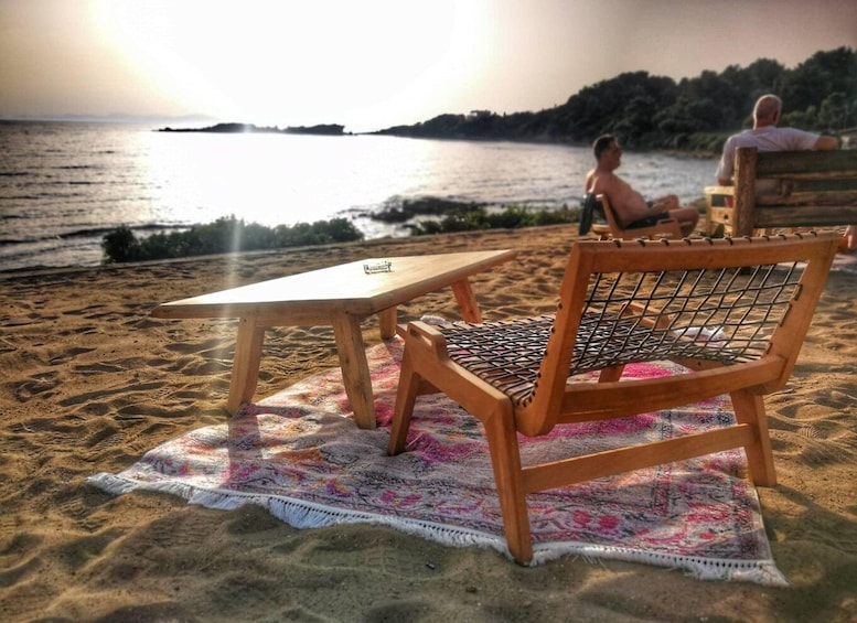 A beach with a wooden chair and table covered by a colorful rug, a person sitting on a bench, and a sunset in the background. 4
