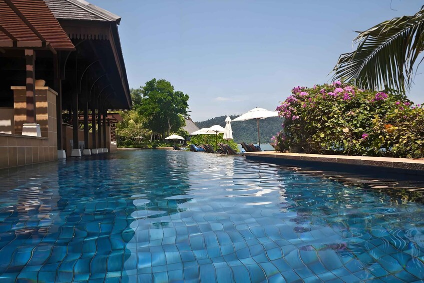 A swimming pool with tiled flooring, surrounded by palm trees and a building with a tiled roof. 5