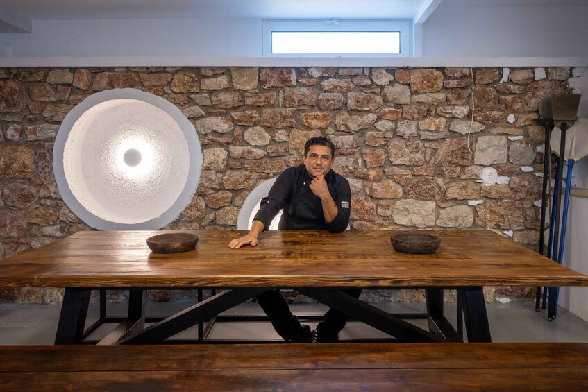 A person seated at a wooden table with a stone wall background and a large circular light fixture. 3