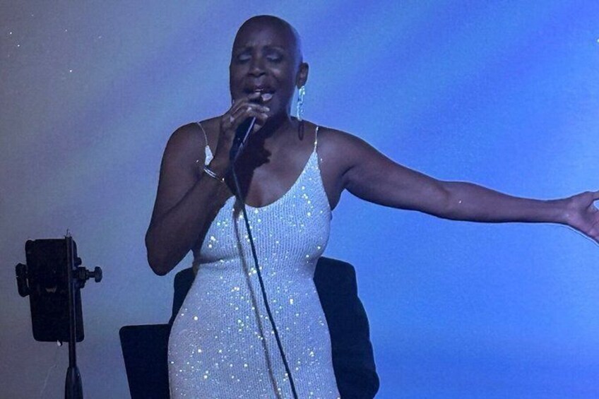 A person in a sparkly dress on stage with a microphone, a camera, and a blue background. 3