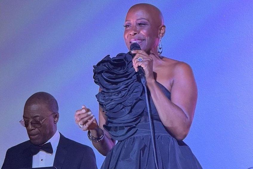 A person holding a microphone, wearing a black dress with ruffles, and standing in front of a blue background. 5
