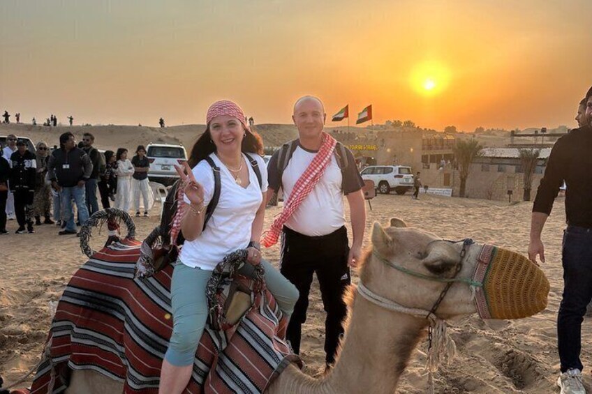 A camel with a rider and a person walking beside it, set against a desert landscape with a sunset. 2