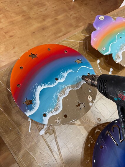 A colorful painted object with a wave design and holes, being worked on with a heat gun. 3