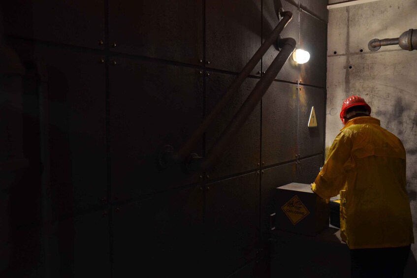 A person in a yellow jacket and red helmet working near a wall with a warning sign. 5