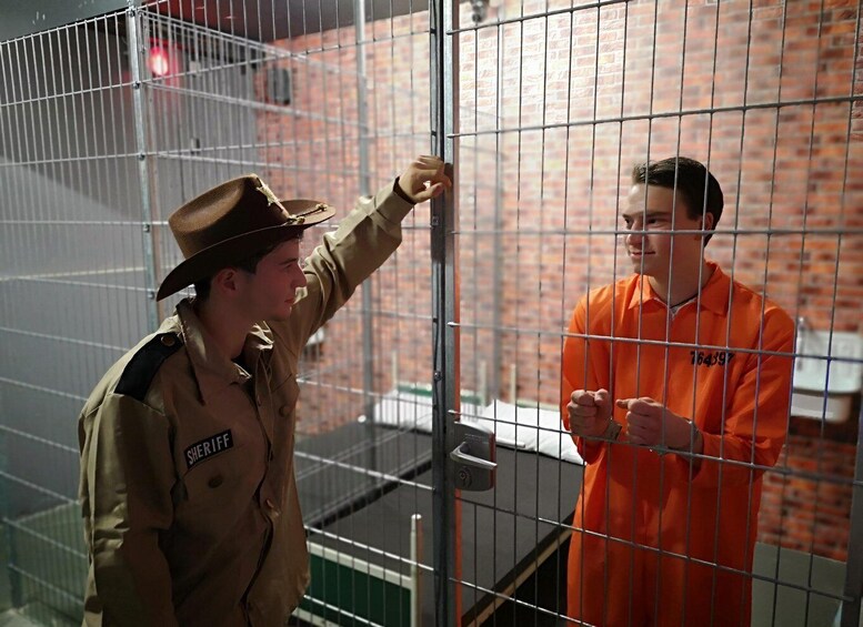 A person in a brown uniform with a hat and another in an orange jumpsuit standing in front of a metal fence. 3