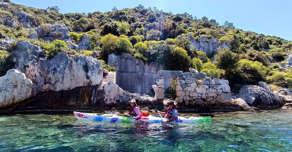 Two people kayaking on a clear waterway with rocky cliffs and greenery in the background. 3