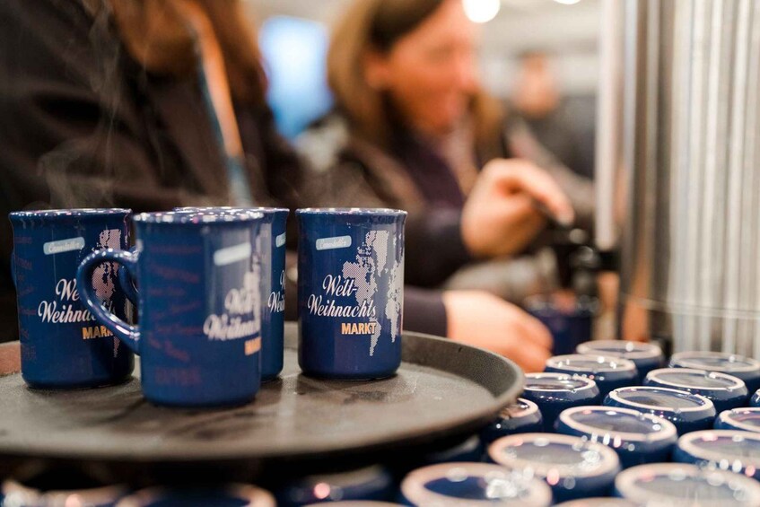 A tray with three blue mugs labeled 'Weihnachts Markt' and a blurred background of people. 2