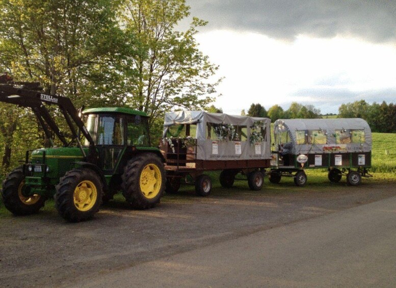 A green tractor with a trailer attached, parked on a road with trees in the background. 1