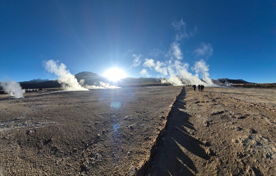 A volcanic landscape with steam vents, a clear sky, and a group of people walking along a path. 3