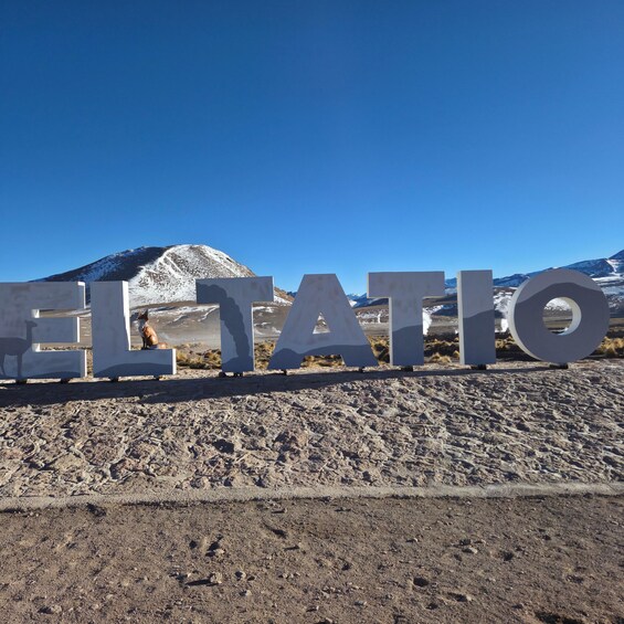 Large metallic letters spelling 'EL TATIO' on a rocky terrain with a clear blue sky and a mountain in the background. 4