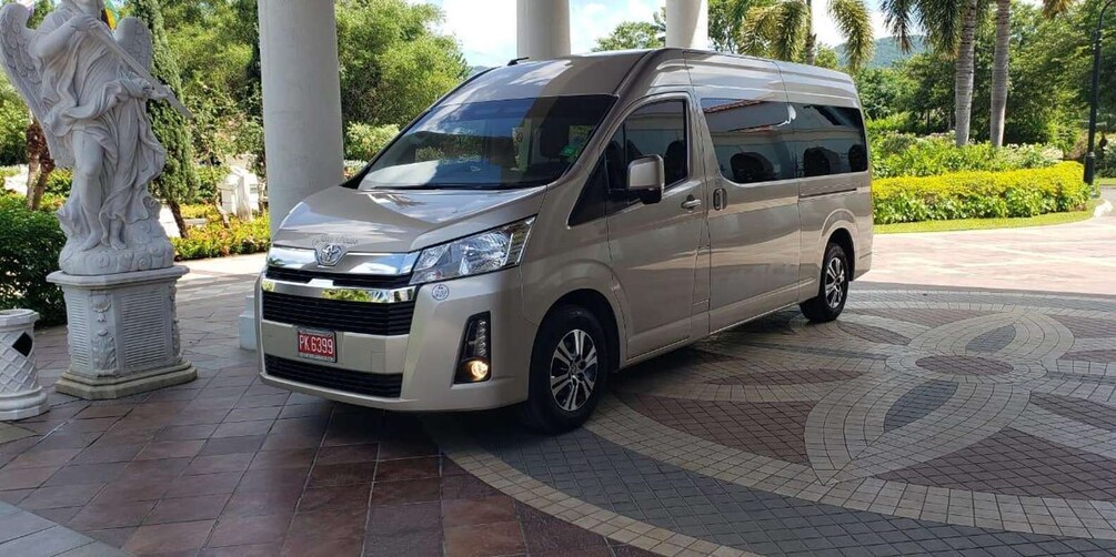 A silver van parked on a patterned pavement with a statue and greenery in the background. 3