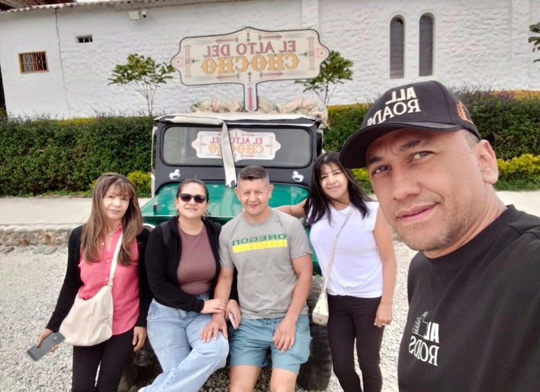 A group of people posing in front of a vintage car and a building with a sign that reads 'EL VOTO DEL CHOCHO'. 2