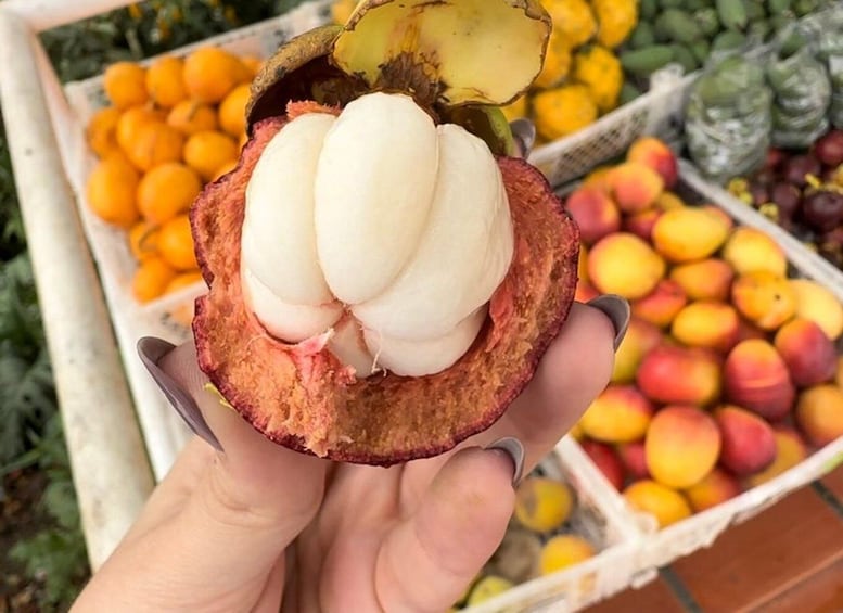 A hand holding a half-open jackfruit with white flesh, surrounded by various fruits at a market. 5