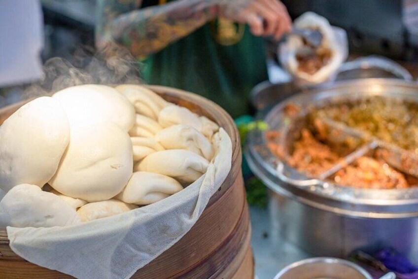 A wooden bowl filled with steamed buns, a person cooking in the background, and a pot of food. 2