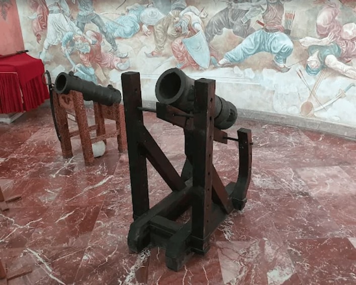 A cannon on a wooden stand with a marble floor and a mural in the background. 2