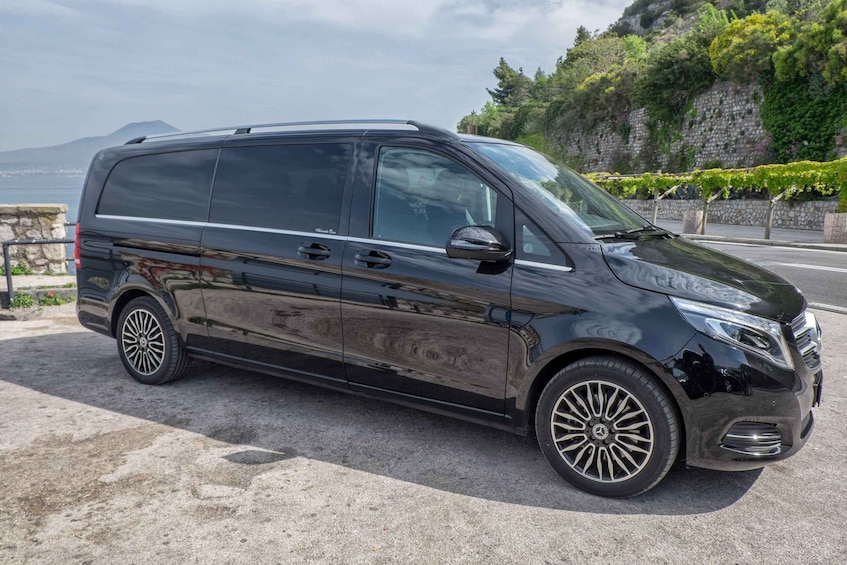 A black van parked on a paved area with a stone wall and greenery in the background. 1