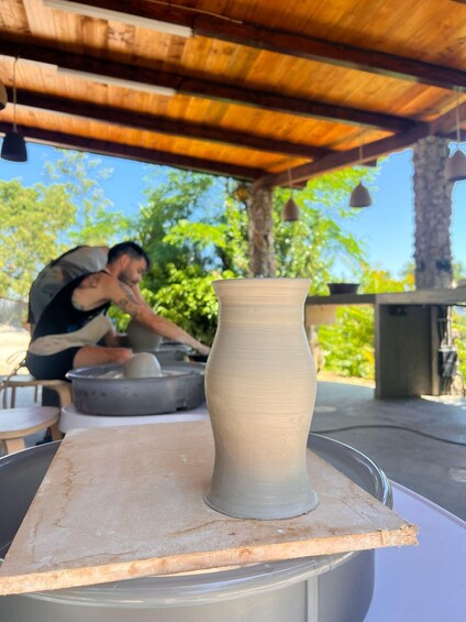A pottery wheel with a partially finished vase, a person working in the background, and a wooden structure overhead. 1