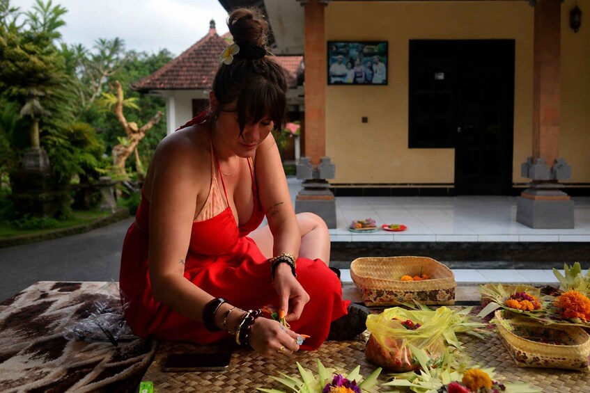 A person in a red dress sitting on a mat, arranging flowers and offerings in front of a building with a tiled roof. 4