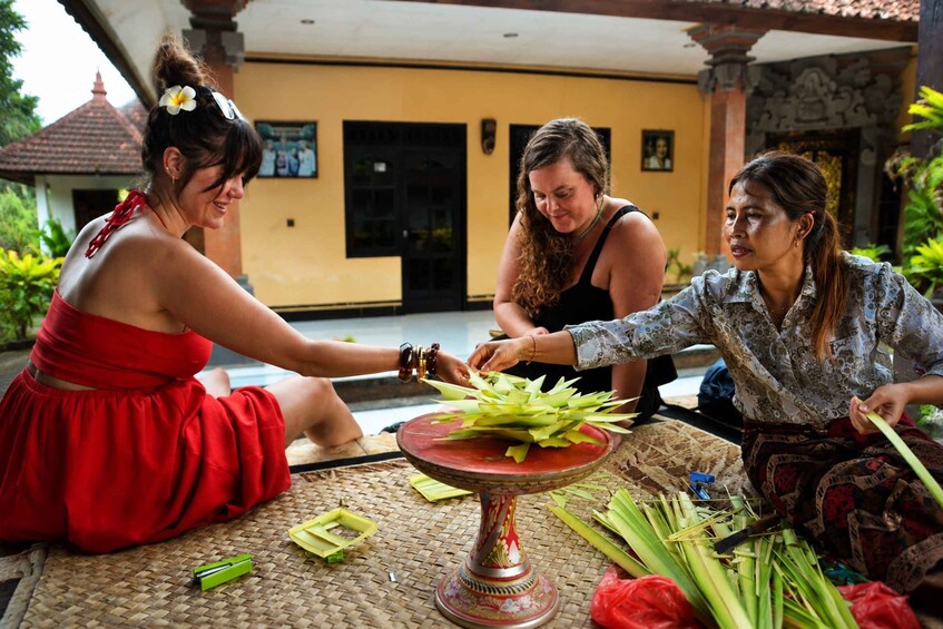 Three women sitting on a woven mat, preparing a ritual with green leaves and a decorated stand. 2
