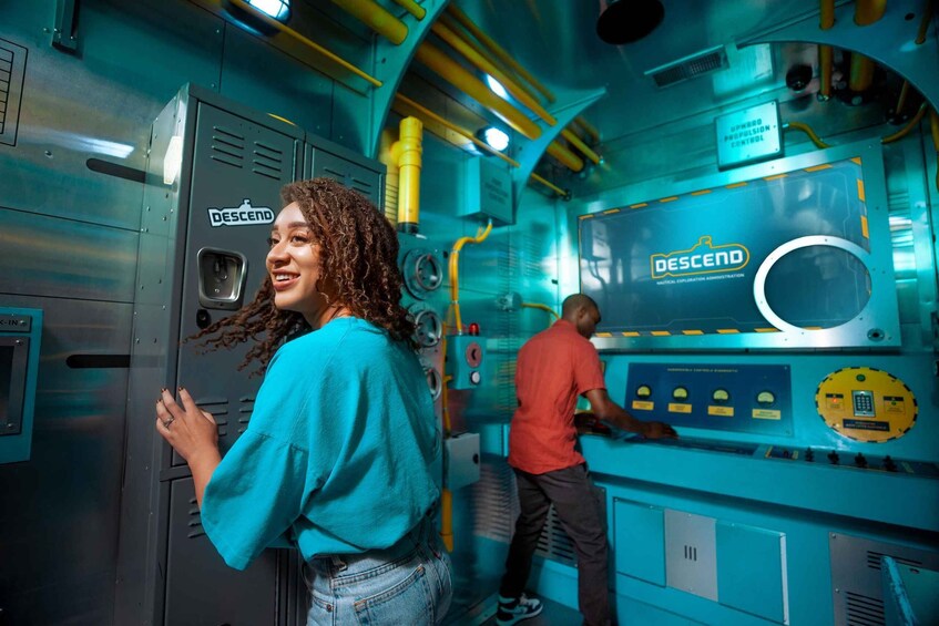 A person in a blue shirt and jeans standing in front of a control panel with the logo 'DESCEND' and a man in a red shirt operating machinery. 4