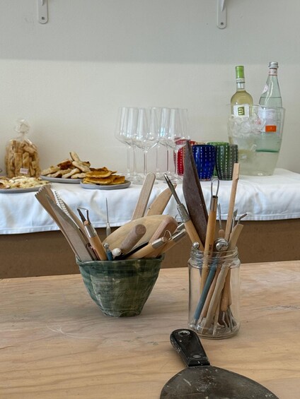 A table with a bowl of wooden utensils, a jar of paintbrushes, a palette knife, and a glass bottle. 2