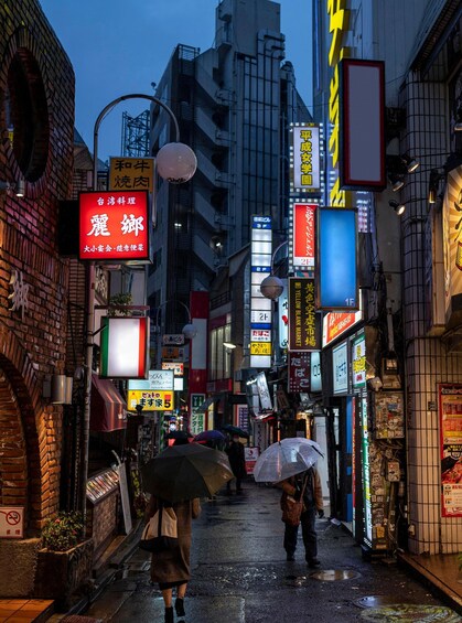A narrow urban street at night with illuminated signs and a wet pavement. 2