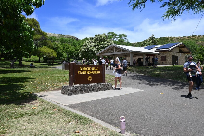A park entrance with a sign for the Diamond Head State Monument, a covered pavilion, and a stone building with solar panels. 1