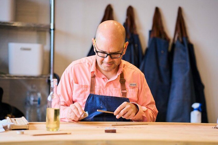 A person wearing a pink shirt and blue apron, working at a wooden table with various items like a glass, a bottle, and a pen. 4