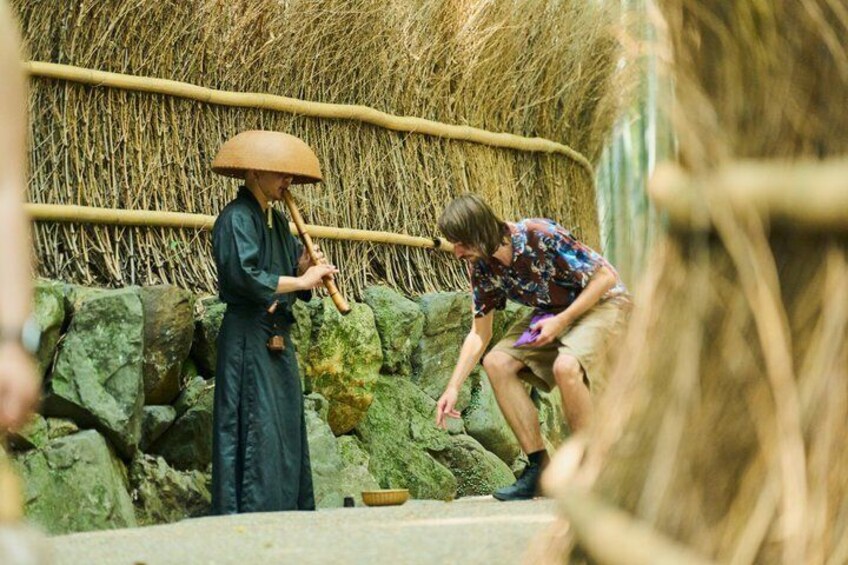 A person playing a traditional bamboo flute, another person crouching, and a straw wall in the background. 4