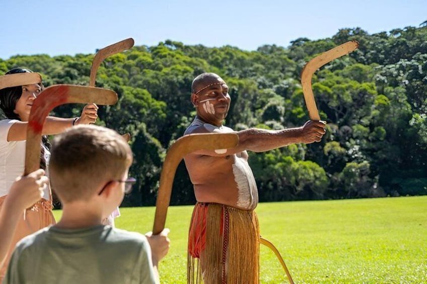 A person in traditional attire holding a long wooden object, with a grassy field and trees in the background. 3