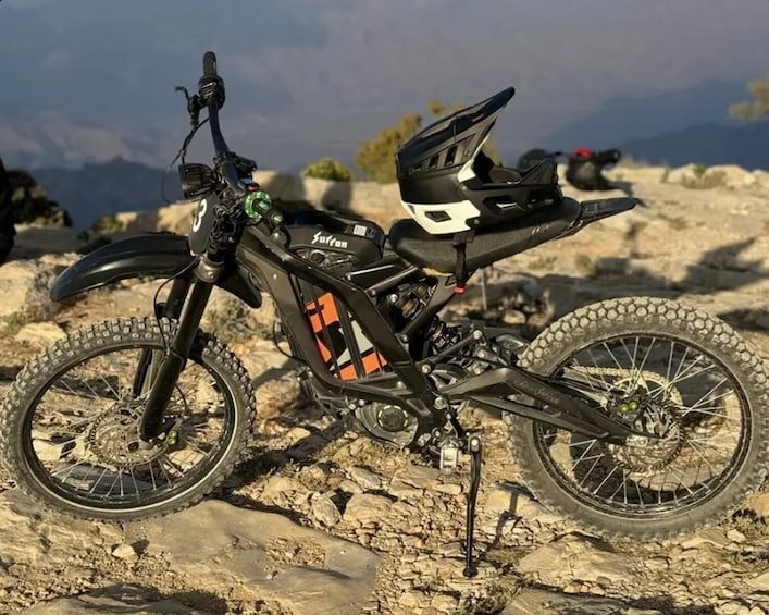 A mountain bike with a black frame and orange accents, equipped with a front suspension fork and a rear suspension system, parked on rocky terrain with a mountainous backdrop. 3