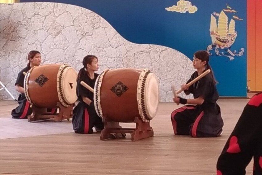 A group of people playing traditional drums in a room with a stone wall and a mural. 3