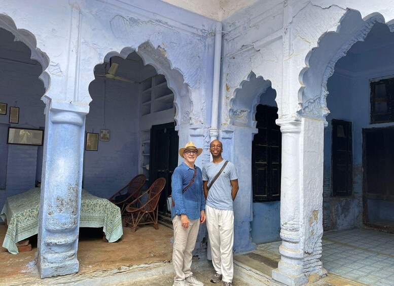 A historic building with white-painted arches and columns, a bed with a patterned bedspread, and two people standing in the doorway. 1