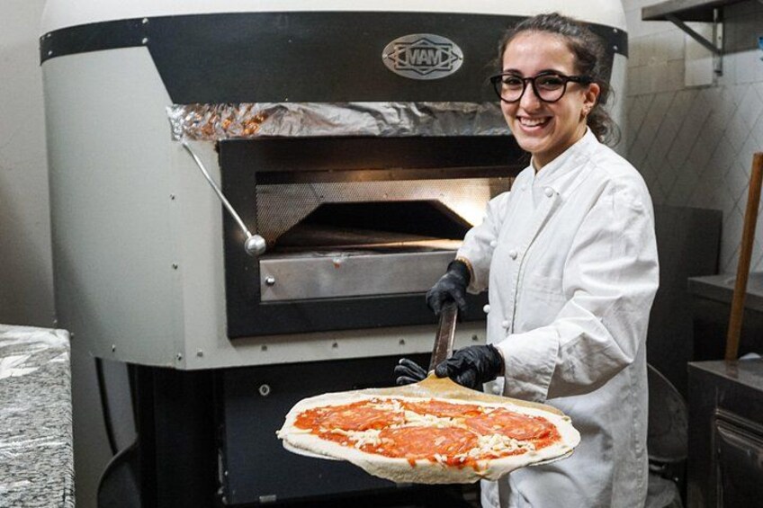A person in a white coat holding a pizza in front of a pizza oven. 1