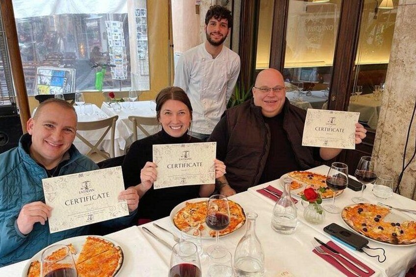 Een groep mensen aan een restauranttafel met pizza, wijnglazen en een certificaat. 2