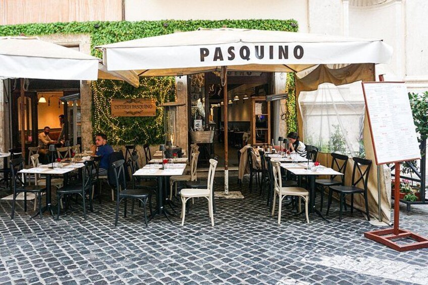 Een terras genaamd PASQUINO met tafels en stoelen, een geplaveid voetpad en groen op de achtergrond. 3