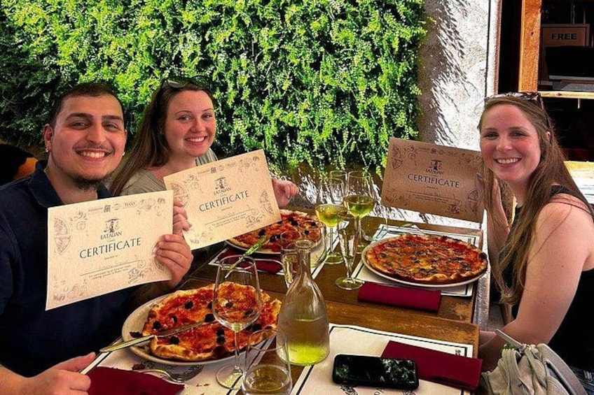 Een groep mensen houdt diploma's vast in een restaurant, met pizza en wijnkaraffen op tafel. 4